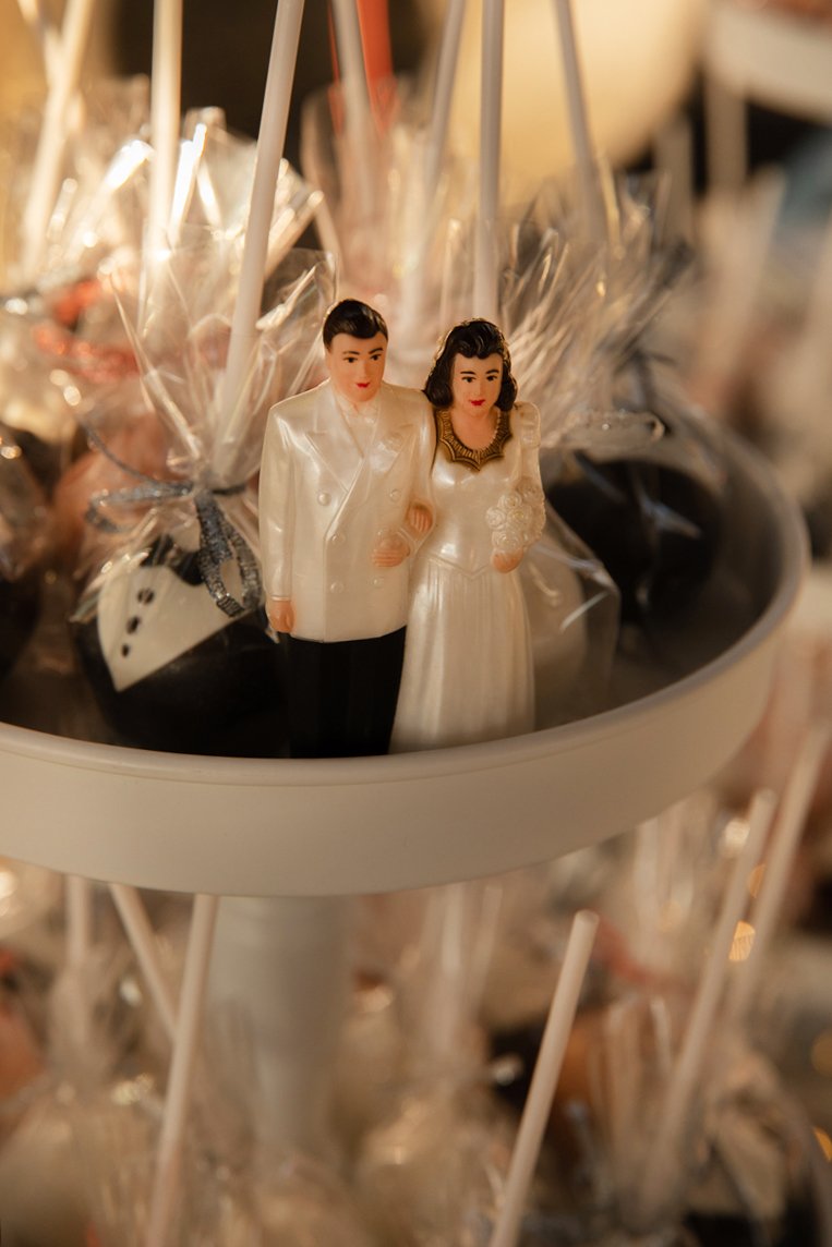 Miniature figurines of a man and woman dressed in wedding attire, standing among wrapped lollipops or candy sticks in a display.
