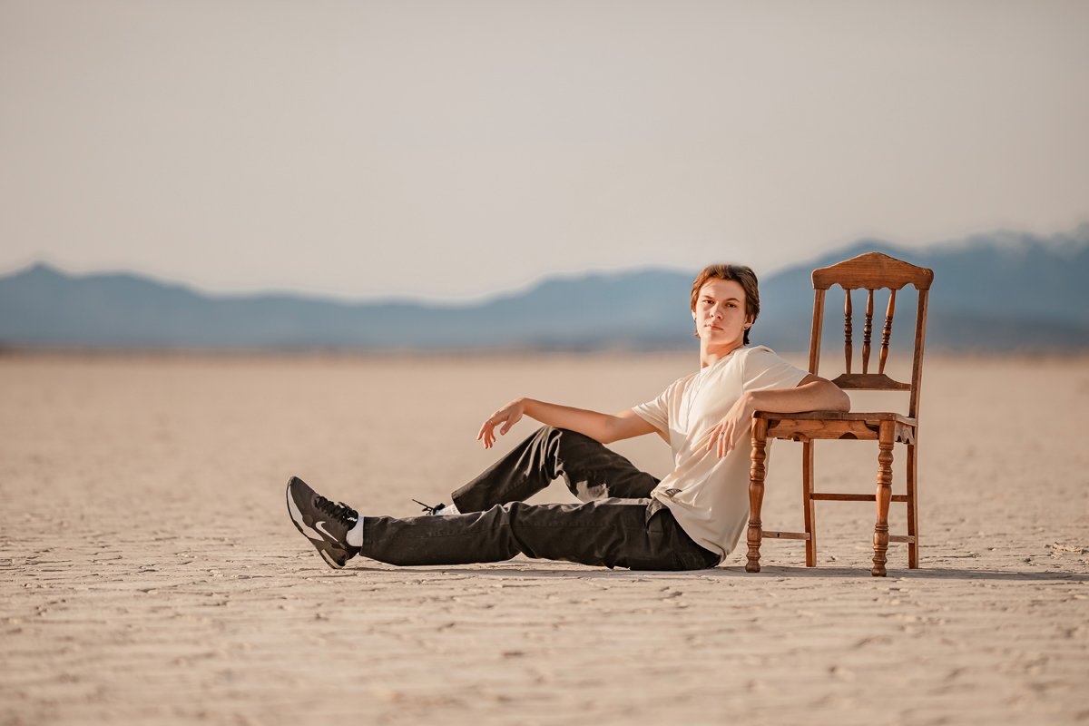 A young man sitting on dry desert ground next to a wooden chair, with mountains in the background and a clear sky.