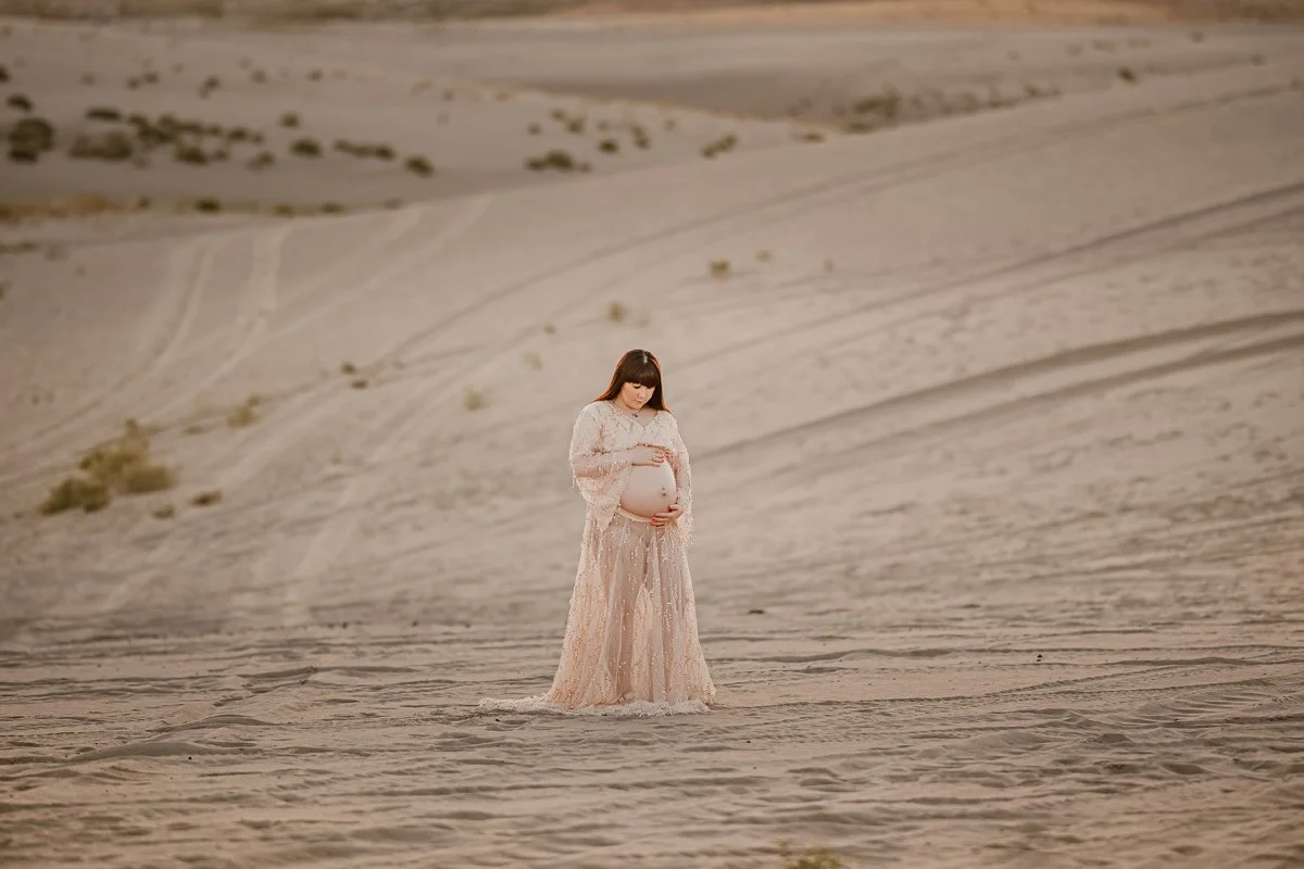 Pregnant woman in a long lace dress standing alone in a barren desert landscape.