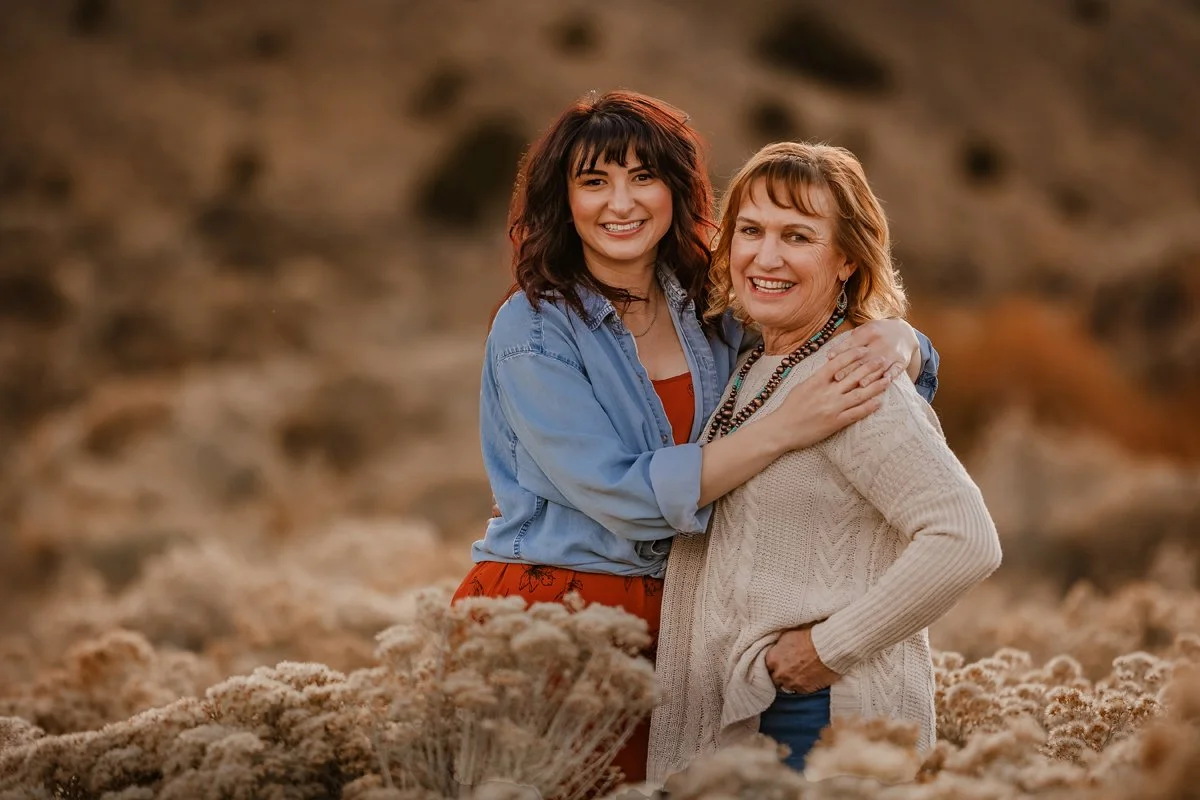 A young woman and an older woman are smiling and hugging outdoors in a field of beige flowers with a blurred background.