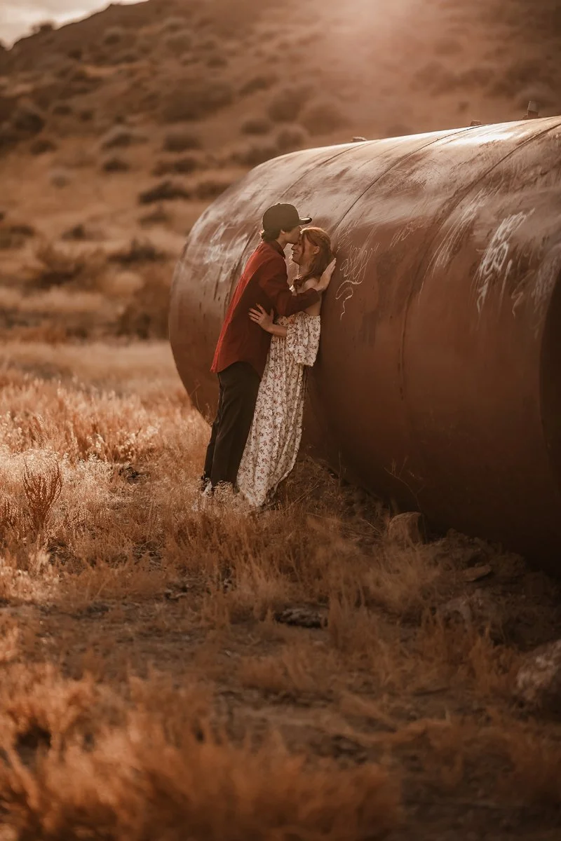 A couple stands close together, nose-to-nose, in front of a large rusty pipeline in a dry, grassy field during sunset.