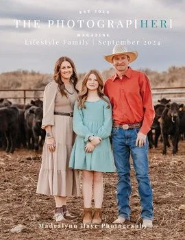 A family of three standing outdoors in a rural landscape with cattle in the background. The woman is wearing a beige dress, the girl is in a blue dress, and the man is in a red shirt and cowboy hat.