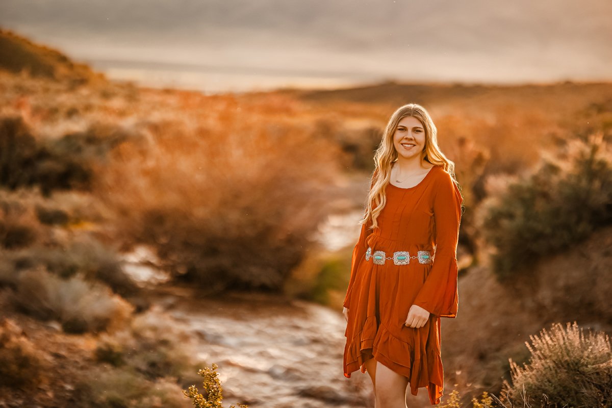 A young woman with long blonde hair standing outdoors in a field during sunset, wearing an orange dress with a decorative belt and smiling at the camera.