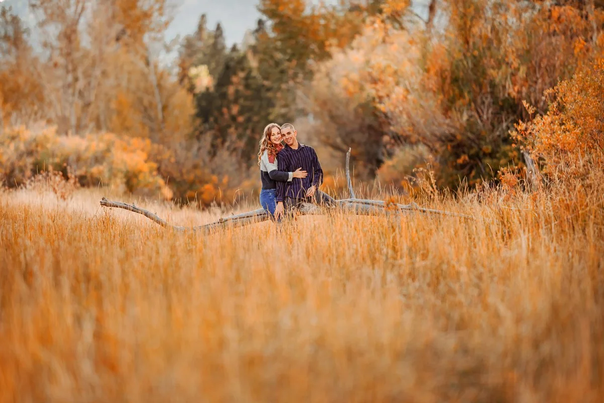 A couple stands in a field of tall, golden grass surrounded by fall foliage with orange and yellow leaves, smiling and embracing.