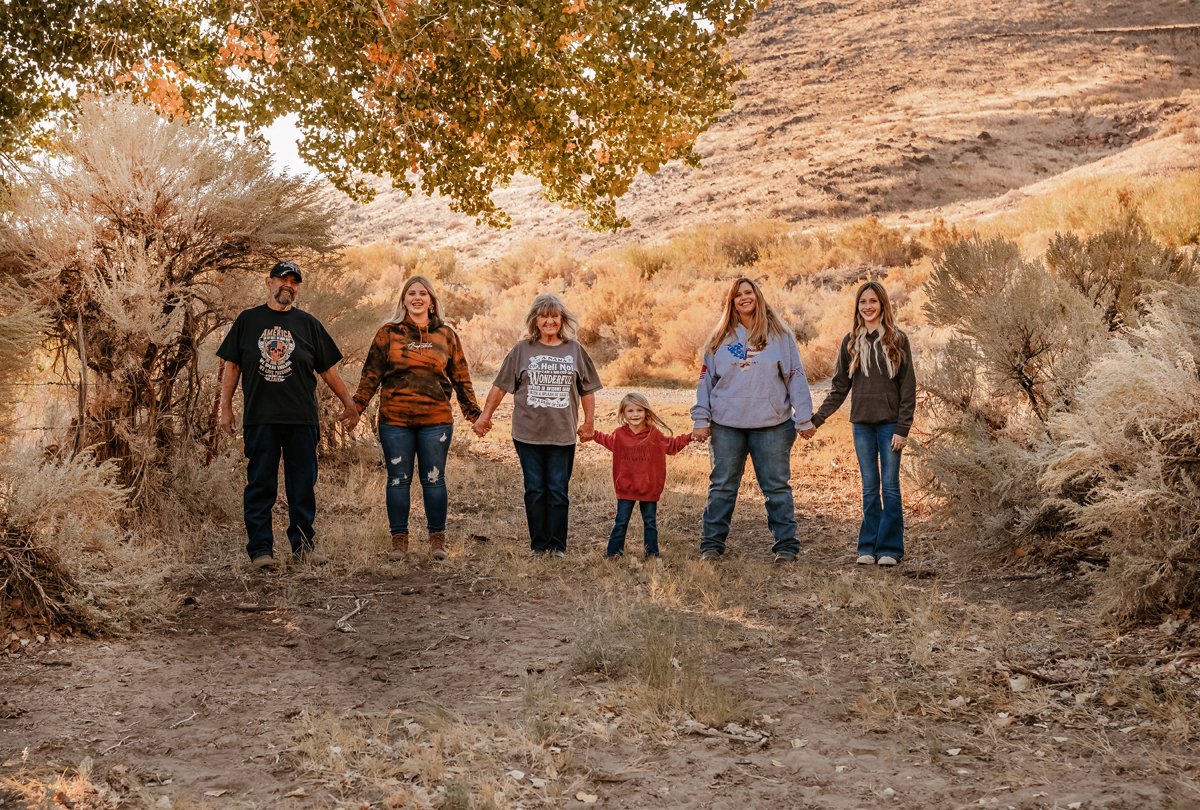 Family of six standing in a line outdoors, holding hands, in a natural landscape with trees and rocky terrain, during a golden hour with warm lighting.