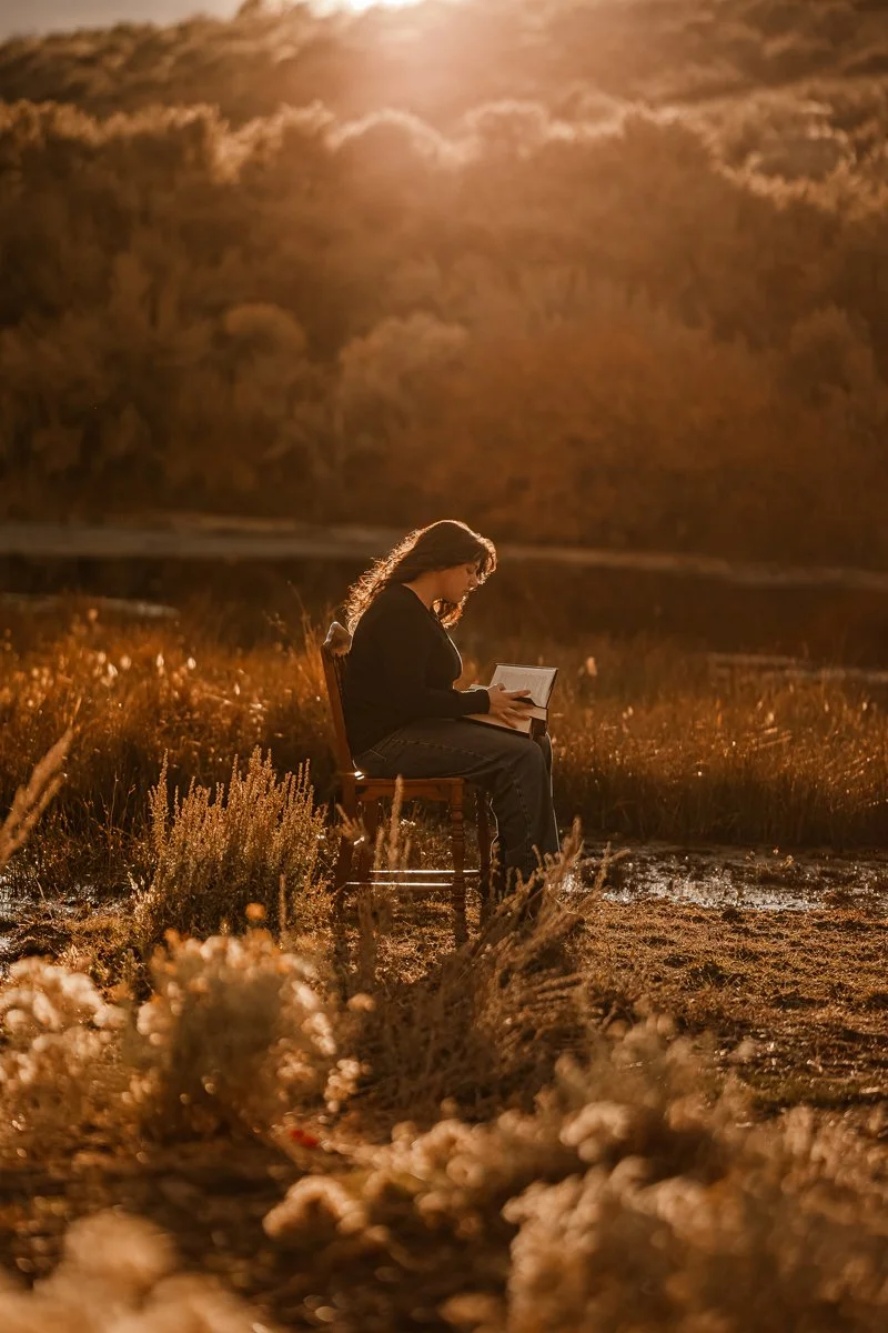 A woman sitting on a chair in a field reading a book during sunset or sunrise, with warm golden light illuminating the scene and a dramatic sky in the background.
