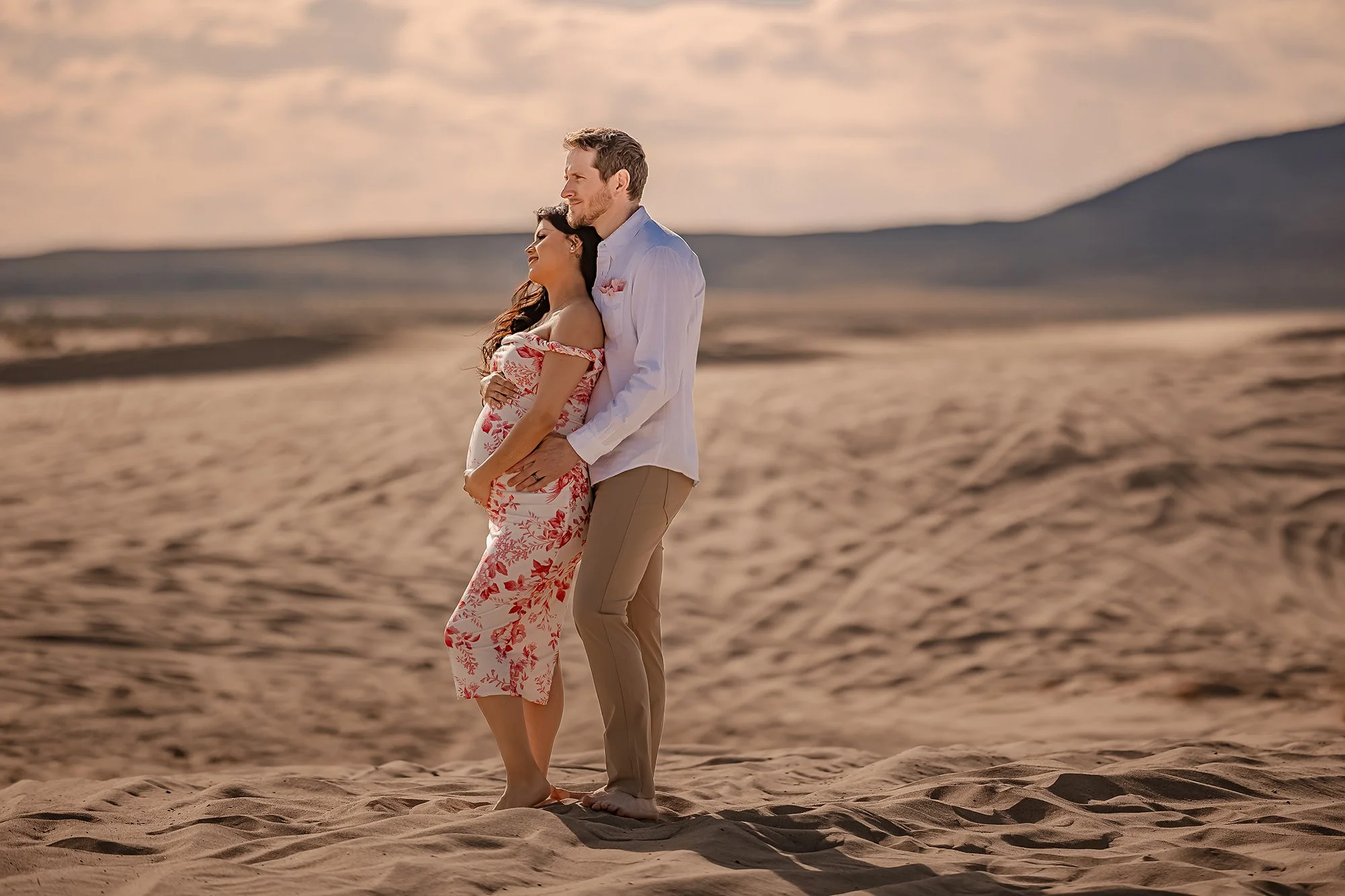 A couple stands on a sandy beach at sunset, with the woman in a floral off-shoulder dress and the man in a white shirt and beige pants, embracing and looking peaceful.