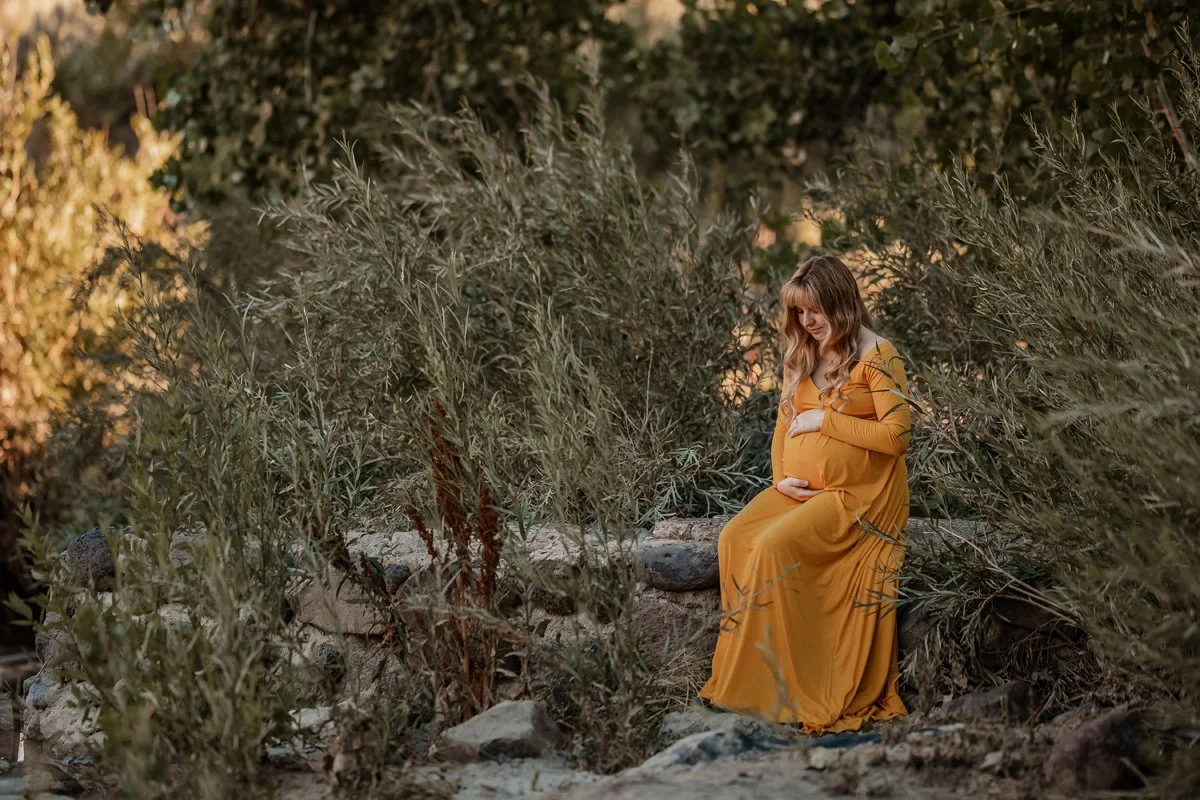 Pregnant woman in a yellow dress sitting on rocks amidst tall bushes and trees in a natural outdoor setting.