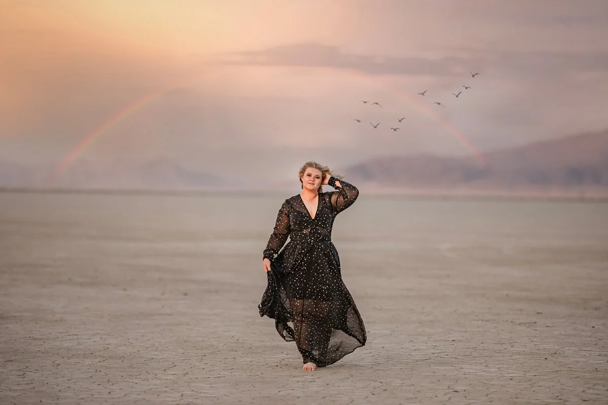 A woman in a black, star-patterned dress walking barefoot on a dry, desert-like landscape under a colorful sky with a rainbow and flying birds.