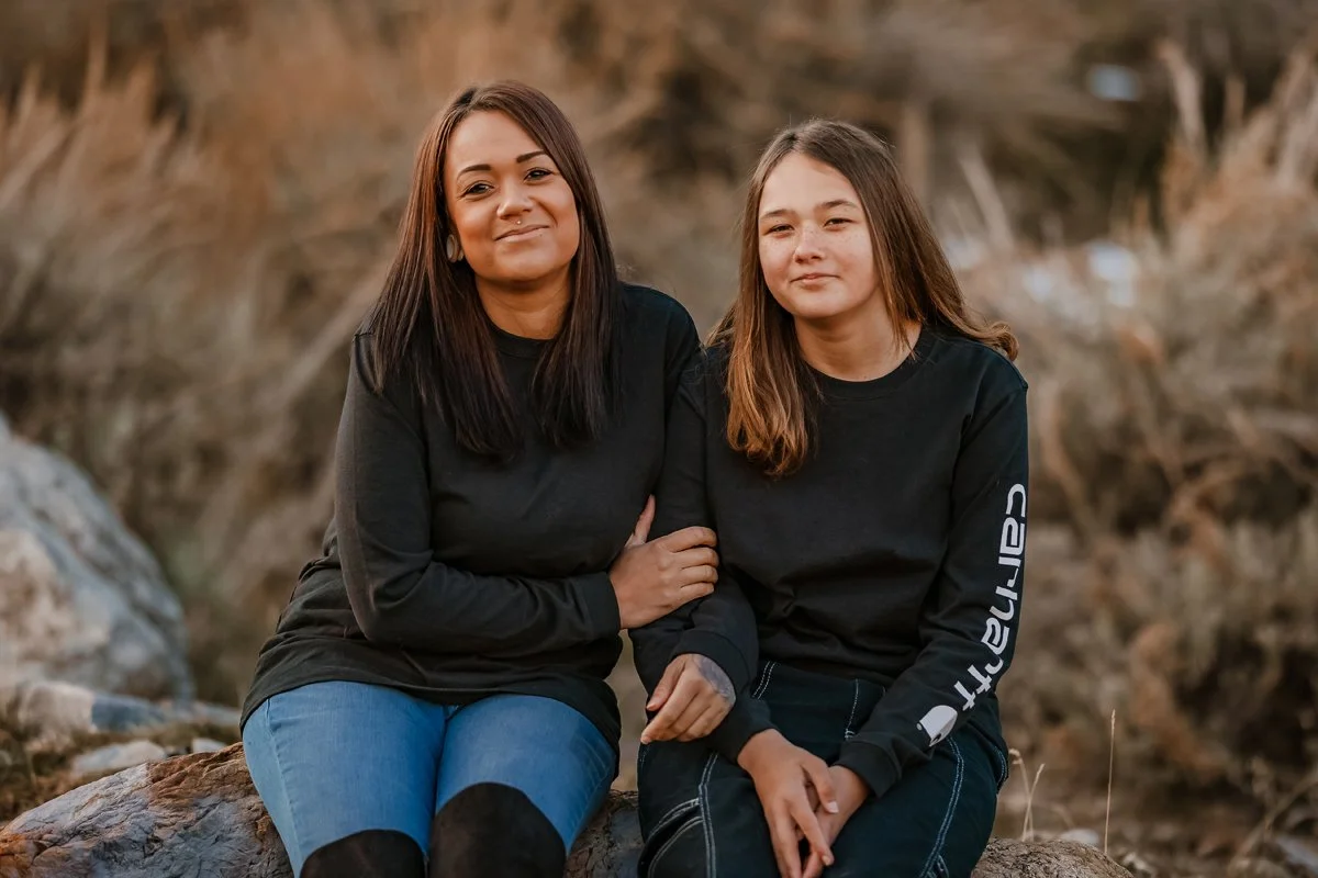 A woman and a girl sitting on rocks outdoors, both wearing black long-sleeve shirts and jeans, posing for a photo with a blurred natural background.