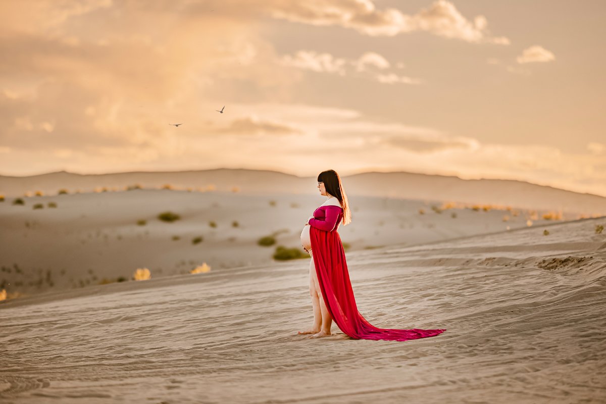 Pregnant woman in a long red dress standing barefoot in a desert landscape during sunset with mountains and sky in the background.