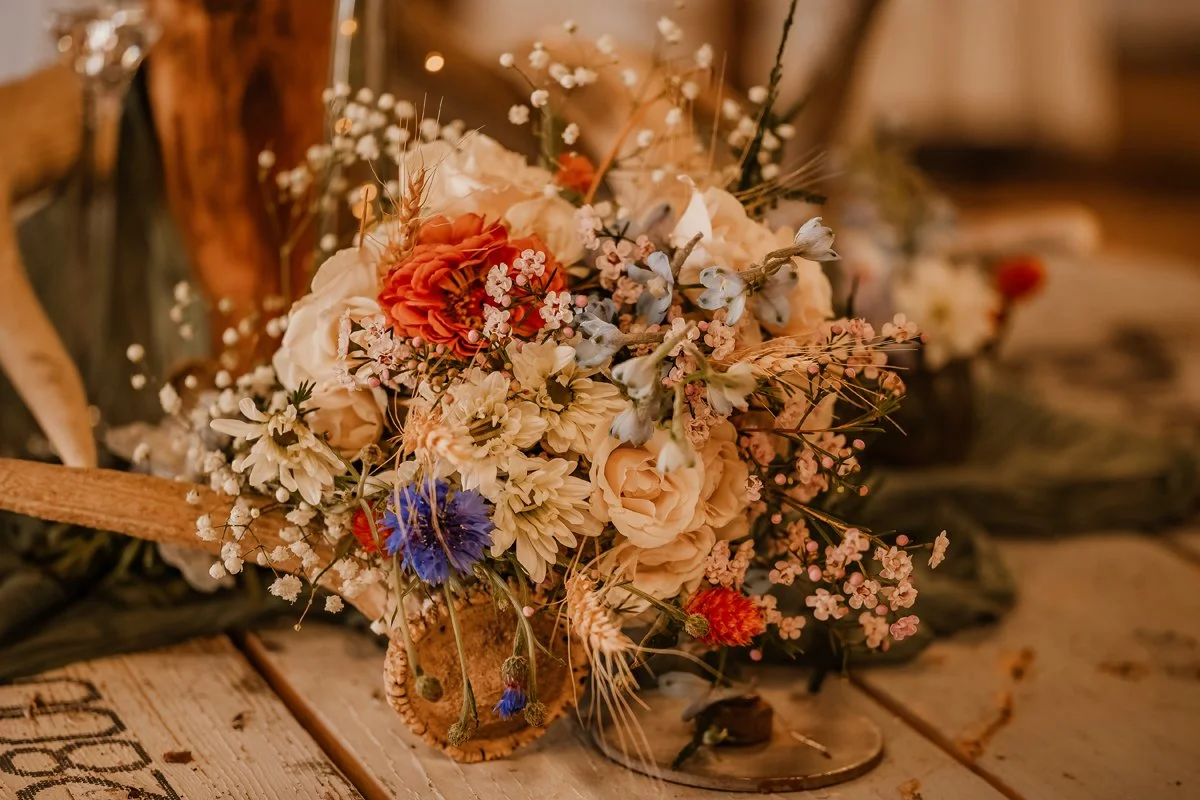 A bouquet of various colored flowers including white, orange, purple, and pink, arranged with greenery and small white filler flowers, placed on a rustic wooden surface.