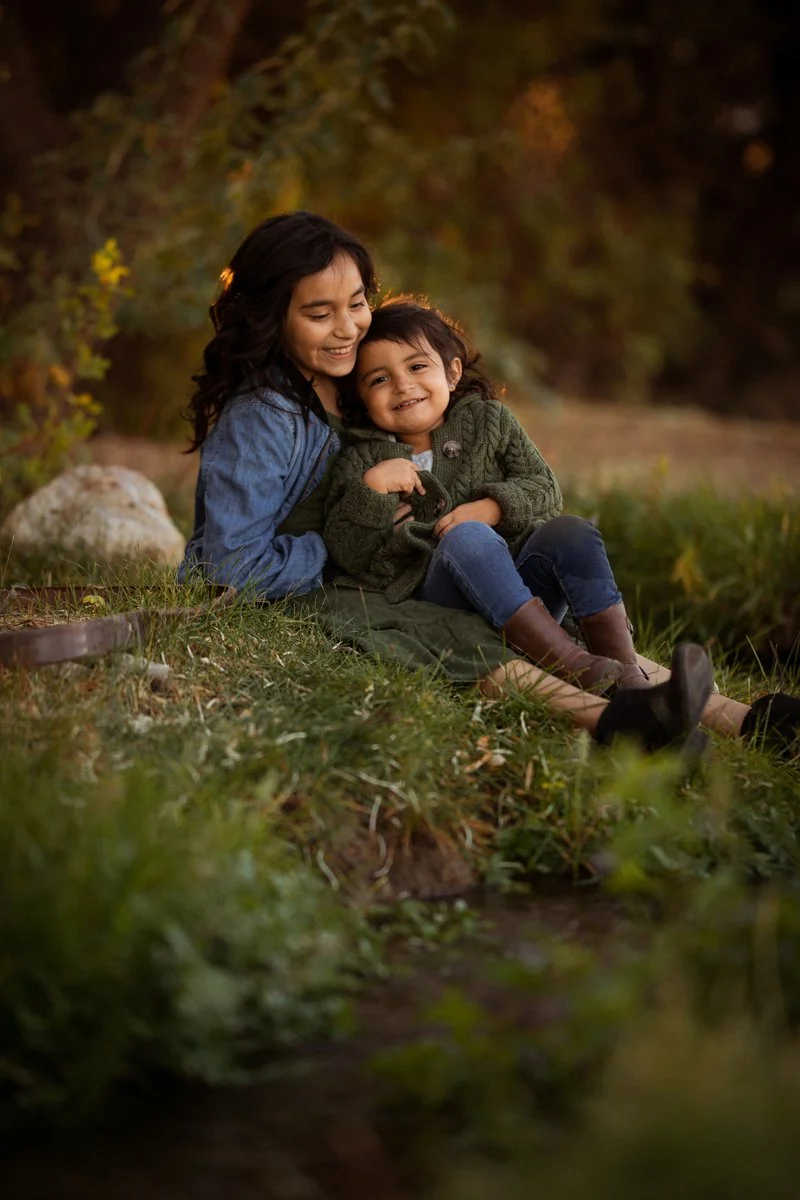 A woman and a young girl sitting together outdoors by a small body of water, smiling and enjoying each other's company during sunset.