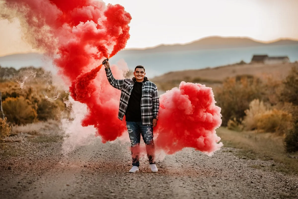 A smiling young man standing outdoors on a dirt path, holding a red smoke flare and surrounded by red smoke clouds, with a rural landscape and distant mountains in the background.