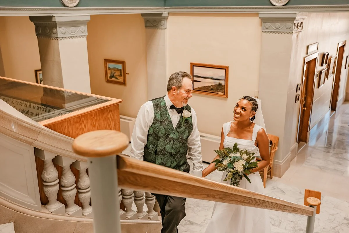 A bride and her father standing on a staircase, holding hands and smiling at each other, with paintings on the wall behind them.