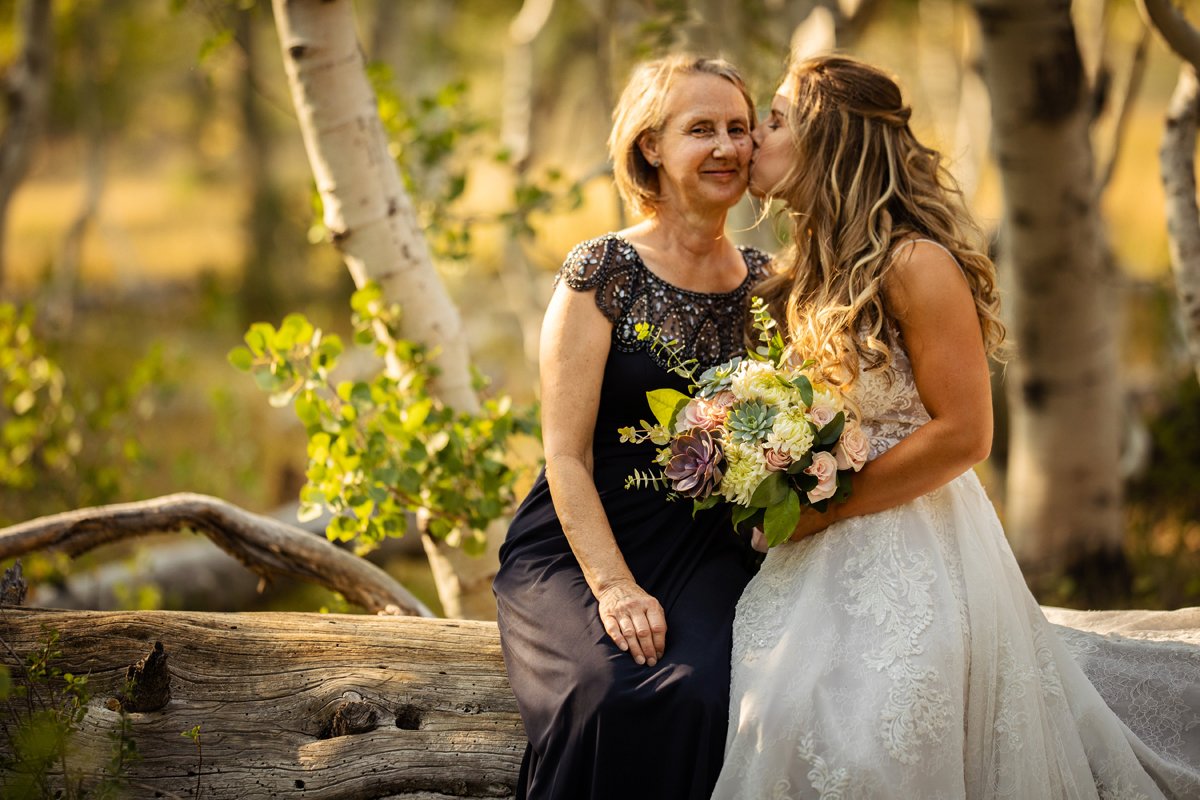 A young bride in a white wedding gown kisses an older woman in a black dress with sequined shoulders on the cheek outdoors during sunset. The woman holds a bouquet of flowers, and they are surrounded by trees and greenery.