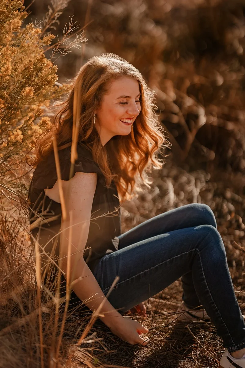 A young woman with long, wavy red hair sitting outdoors in a natural setting, smiling and enjoying the sunlight amidst dry grass and bushes.