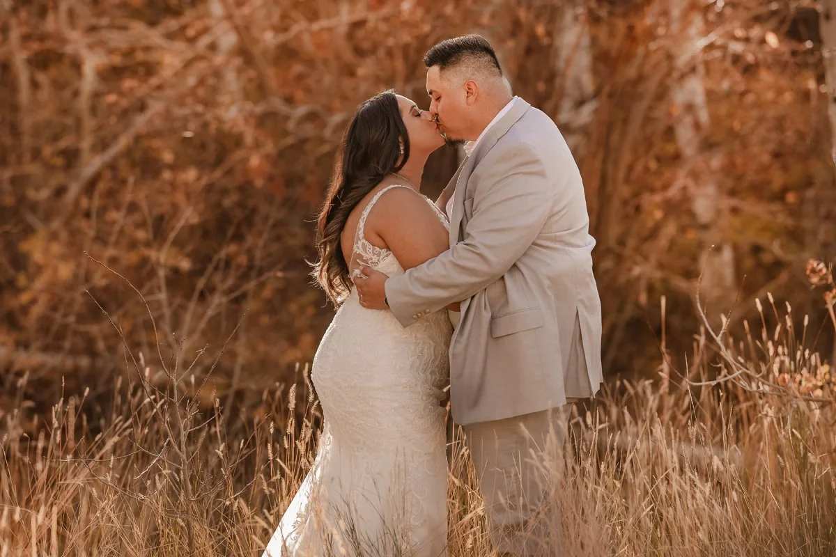 A bride and groom sharing a kiss outdoors in a field surrounded by autumn trees.
