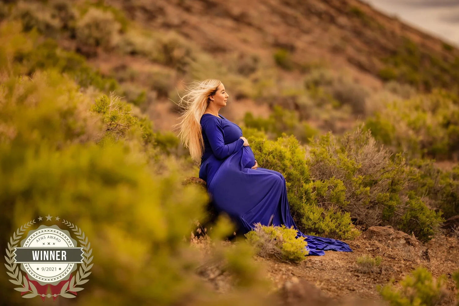 A pregnant woman in a blue dress sitting outdoors on a dirt path surrounded by green bushes and shrubs, with a mountainous background.