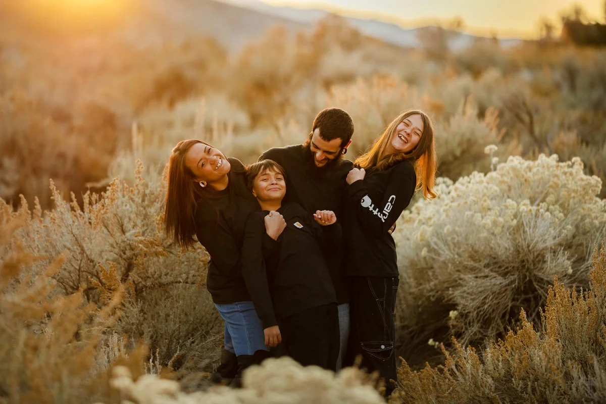 A family of four hugging and smiling together outdoors in a desert-like landscape during sunset.