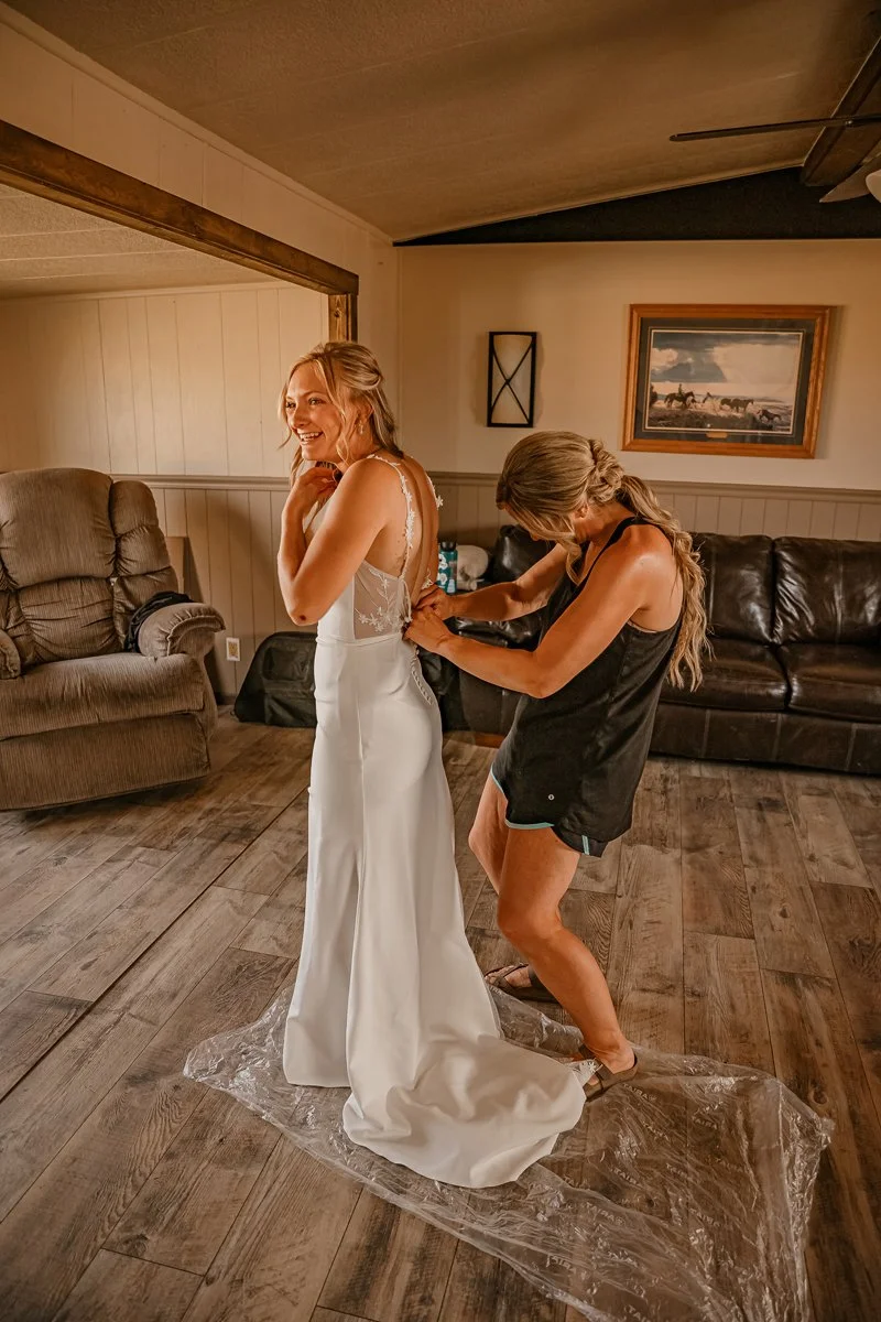 A woman in a wedding dress is smiling while another woman helps her with the dress in a living room.