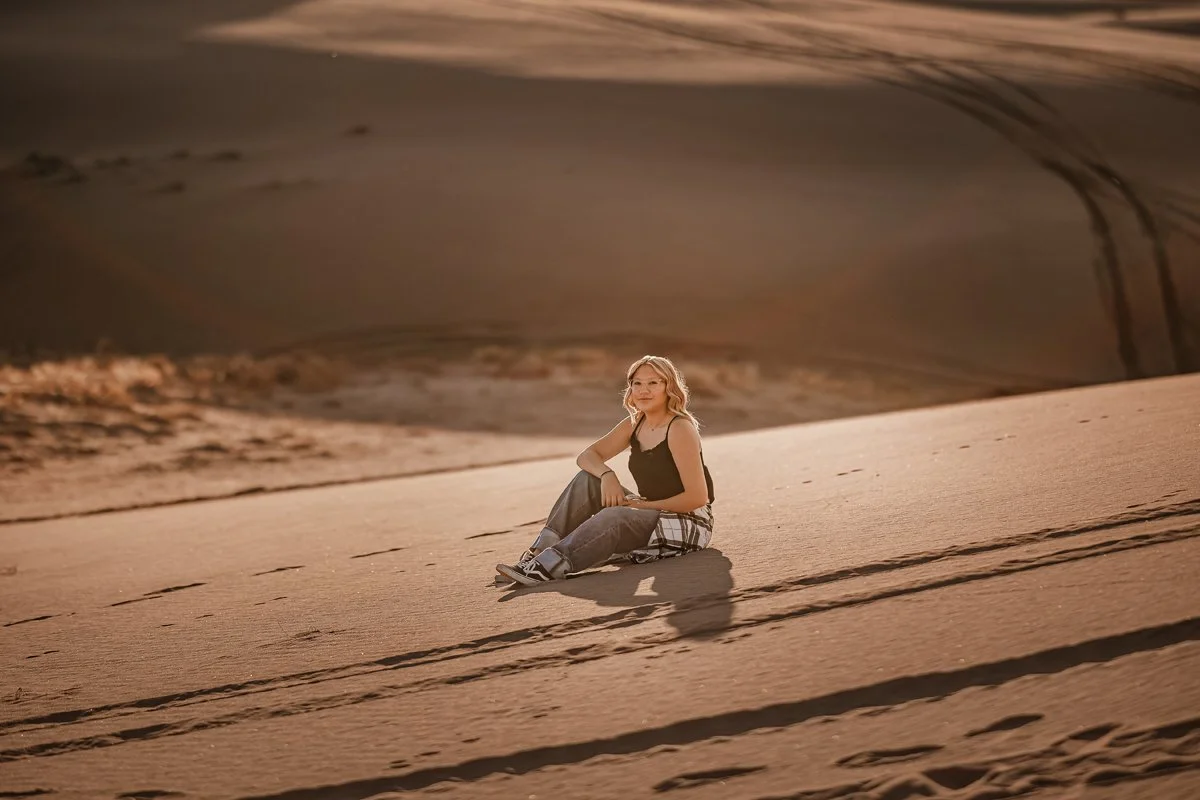 A woman sitting on a sand dune in a desert landscape during sunset.