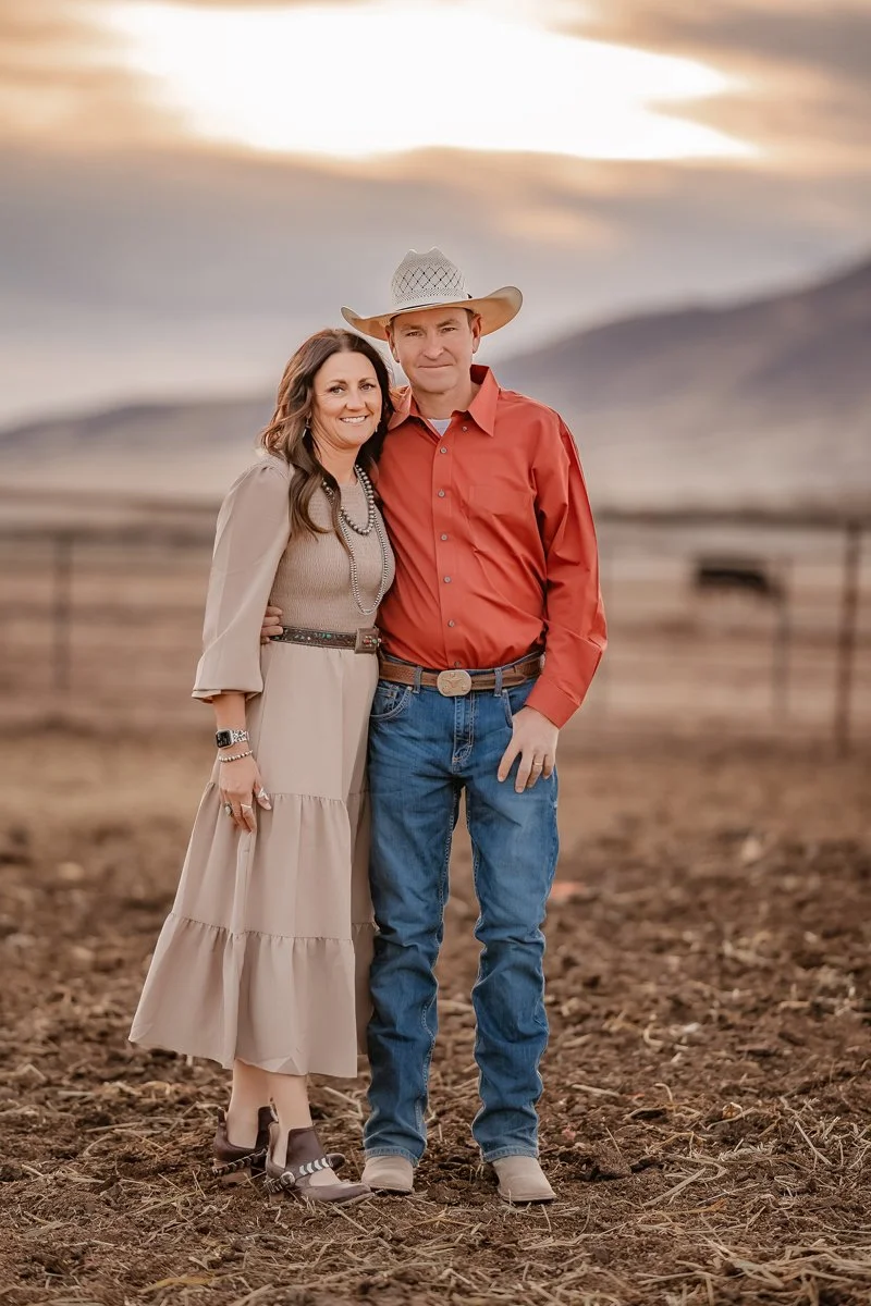 A couple stands outdoors in a rural area during sunset, with a fence and hills in the background. The woman wears a beige dress with jewelry and heels, and the man wears a cowboy hat, red shirt, jeans, and cowboy boots.
