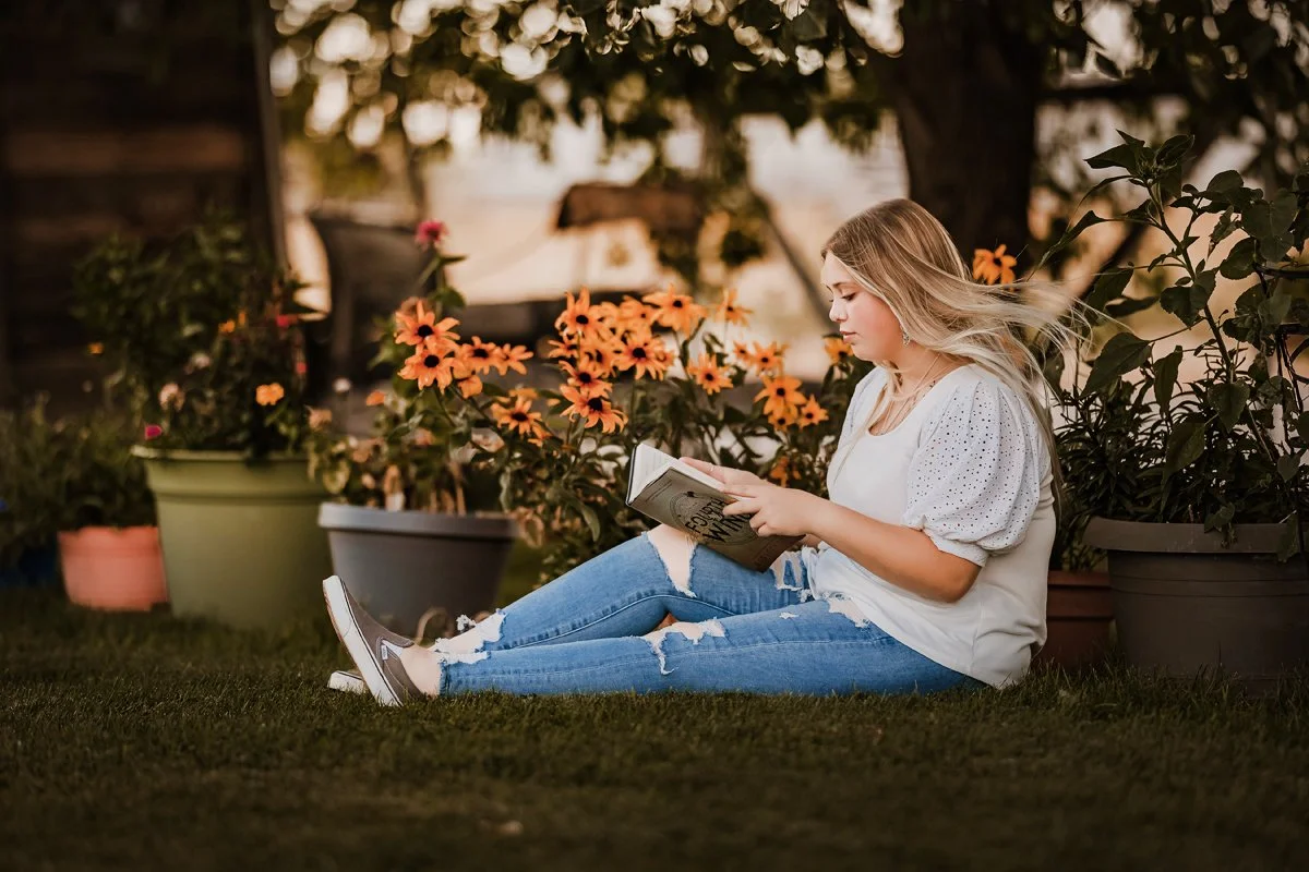 A young woman with blonde hair wearing ripped jeans and a white t-shirt sits on the grass, reading a book, surrounded by colorful flowers in pots, with a large tree and a sunset sky in the background.