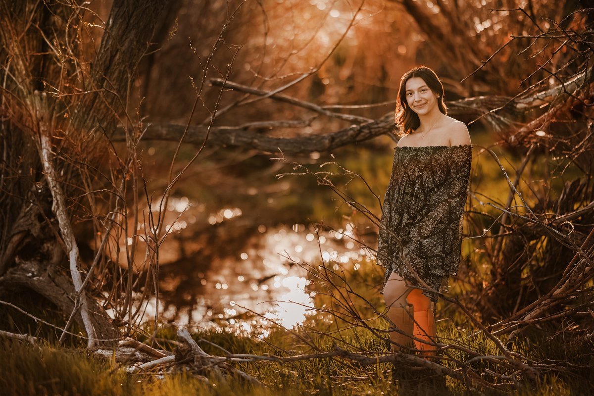 A young woman with dark hair, wearing an off-the-shoulder black dress with a floral pattern, standing outdoors near a tree by a small body of water during sunset or golden hour.