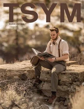 A man sitting outdoors on a large rock, reading a magazine or book, with a blurred natural background.