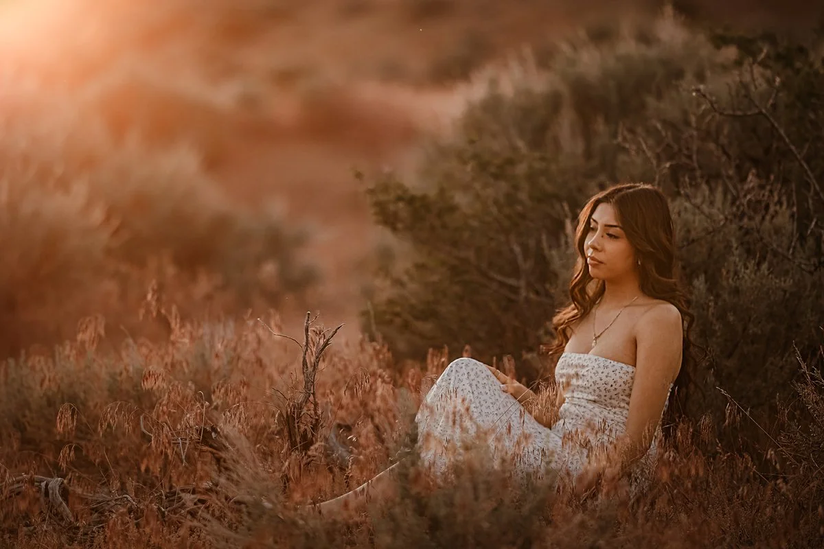 A young woman with long wavy hair sitting in tall, dry grass during sunset in a natural outdoor setting.