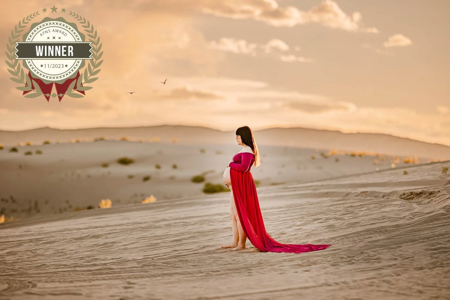 A pregnant woman wearing a long red dress standing barefoot on sand dunes at sunset or sunrise, with a sky filled with clouds and birds flying in the distance.
