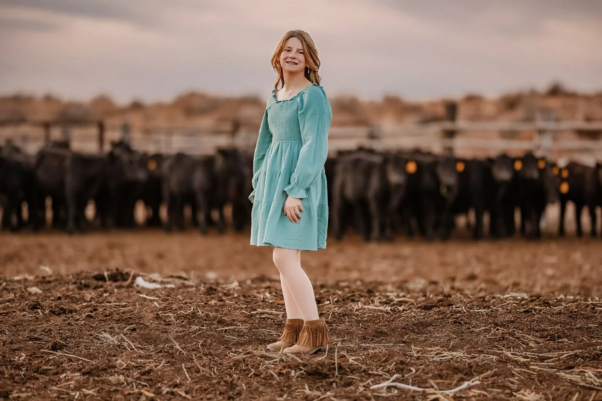 A young woman in a teal dress and tan fringed boots standing on dirt, with cattle behind her in a rural setting at dusk.