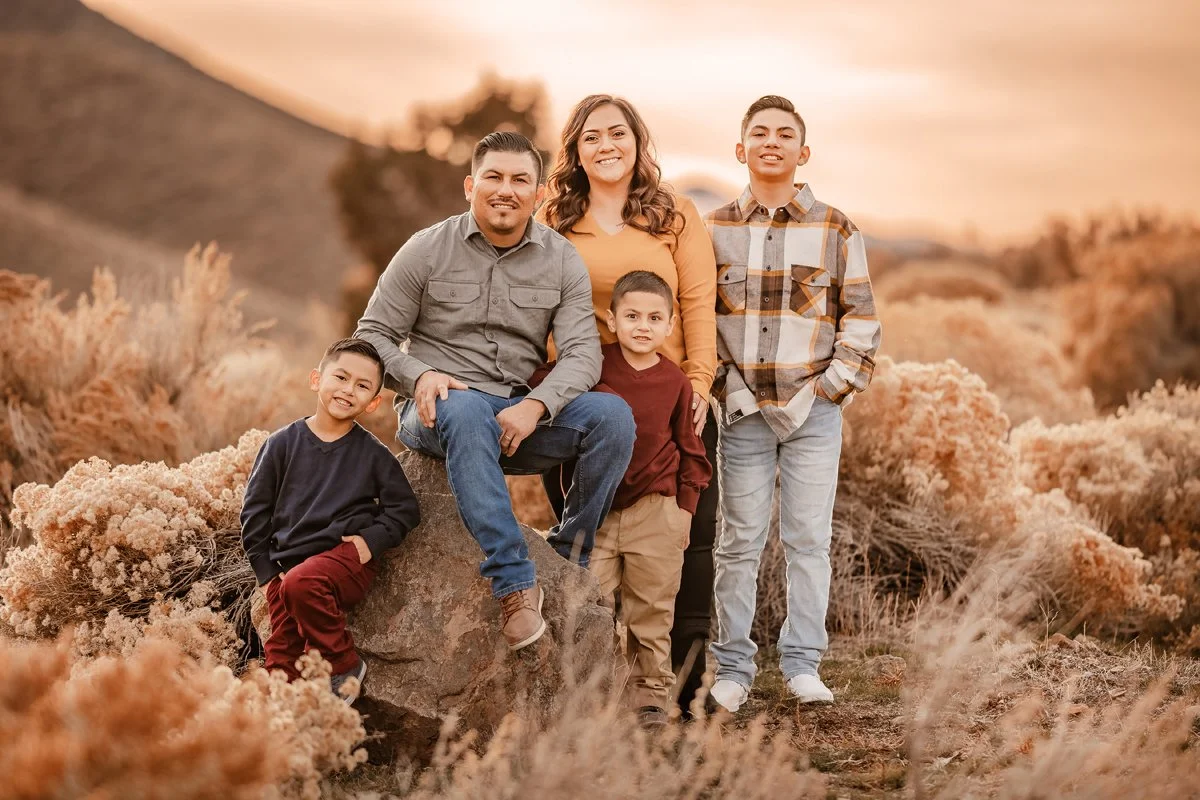 Family of six posing outdoors during sunset, with a landscape of shrubs and hills in the background.
