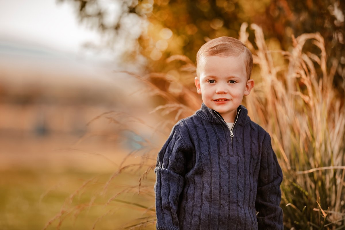 A young boy with short hair, wearing a dark sweater, standing outdoors among tall grass during autumn.