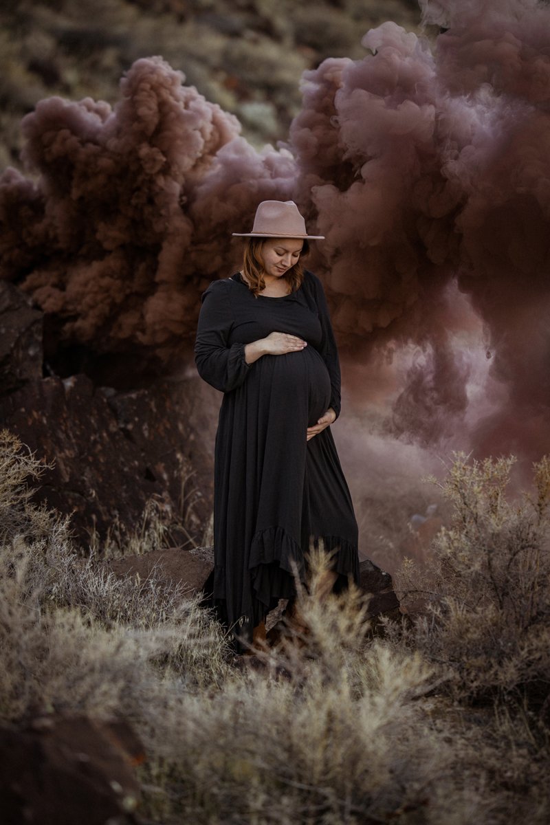 A pregnant woman in a black dress and pink hat stands outdoors in a desert-like area with dry bushes, smiling gently and holding her belly, with large pink smoke clouds billowing in the background.