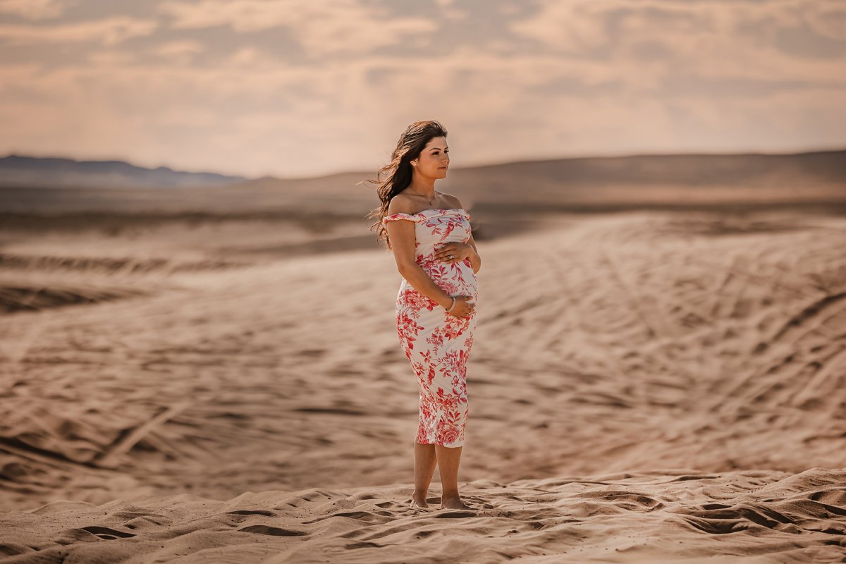 A woman standing barefoot on sandy desert landscape during sunset, wearing a white dress with pink floral patterns, gazing into the distance.