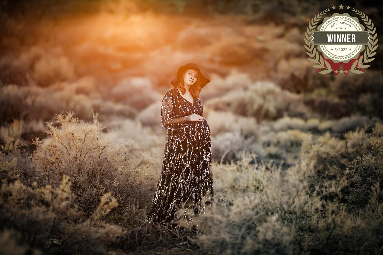 A pregnant woman wearing a long black dress with white patterns and a wide-brimmed black hat stands in a desert landscape with dry bushes and rocks, illuminated by warm sunlight, with a 'winner' badge in the upper right corner.