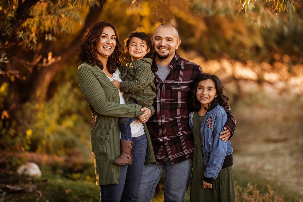 Family of four smiling outdoors with autumn leaves in background, embracing and posing for a photo