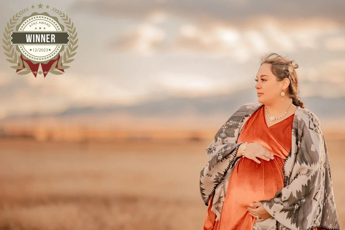 A pregnant woman standing outdoors during sunset, wearing a rust-colored dress and a patterned shawl, with her hand on her belly, looking to the right.