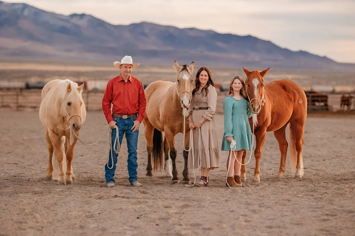 A family with three people and three horses standing in an outdoor arena with mountains in the background.