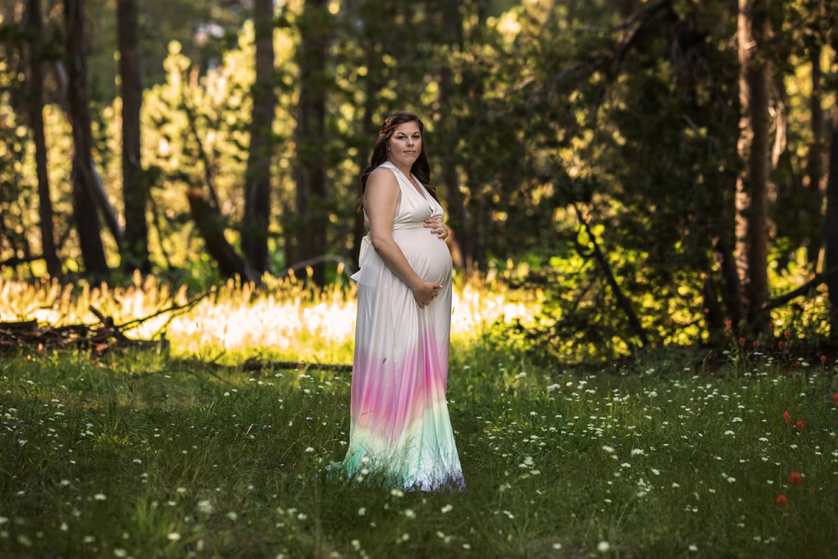 Pregnant woman standing in a forest clearing, wearing a colorful gradient dress, with trees and sunlight in the background.