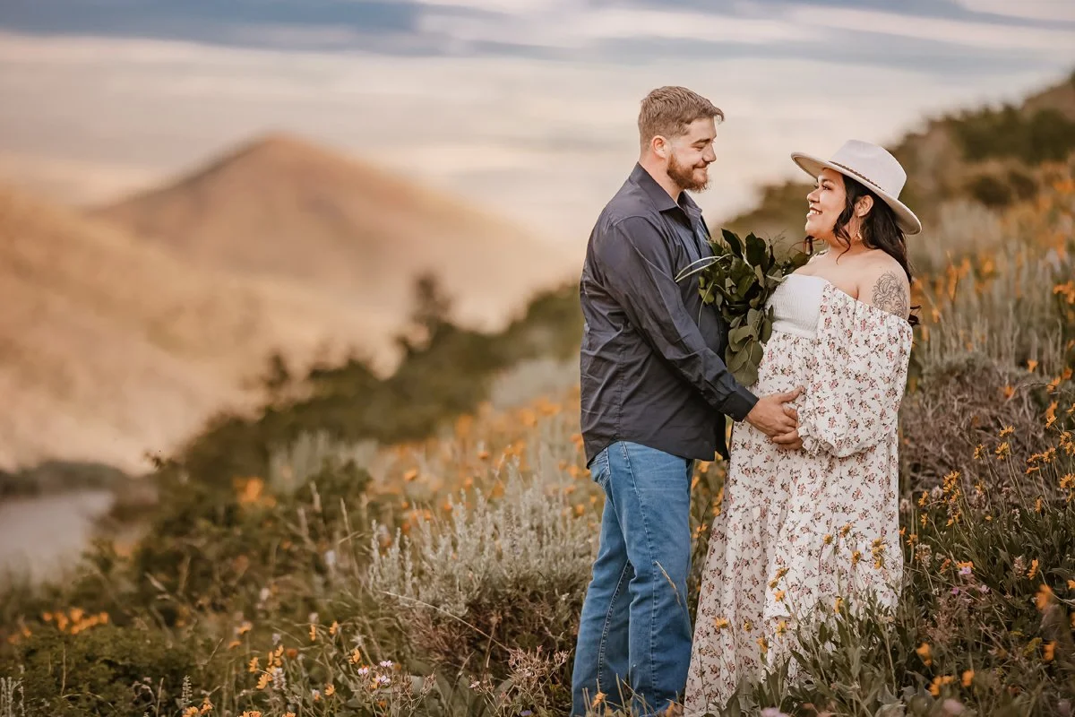 A couple standing in a field on a hillside, exchanging a loving look, with mountains and a cloudy sky in the background.