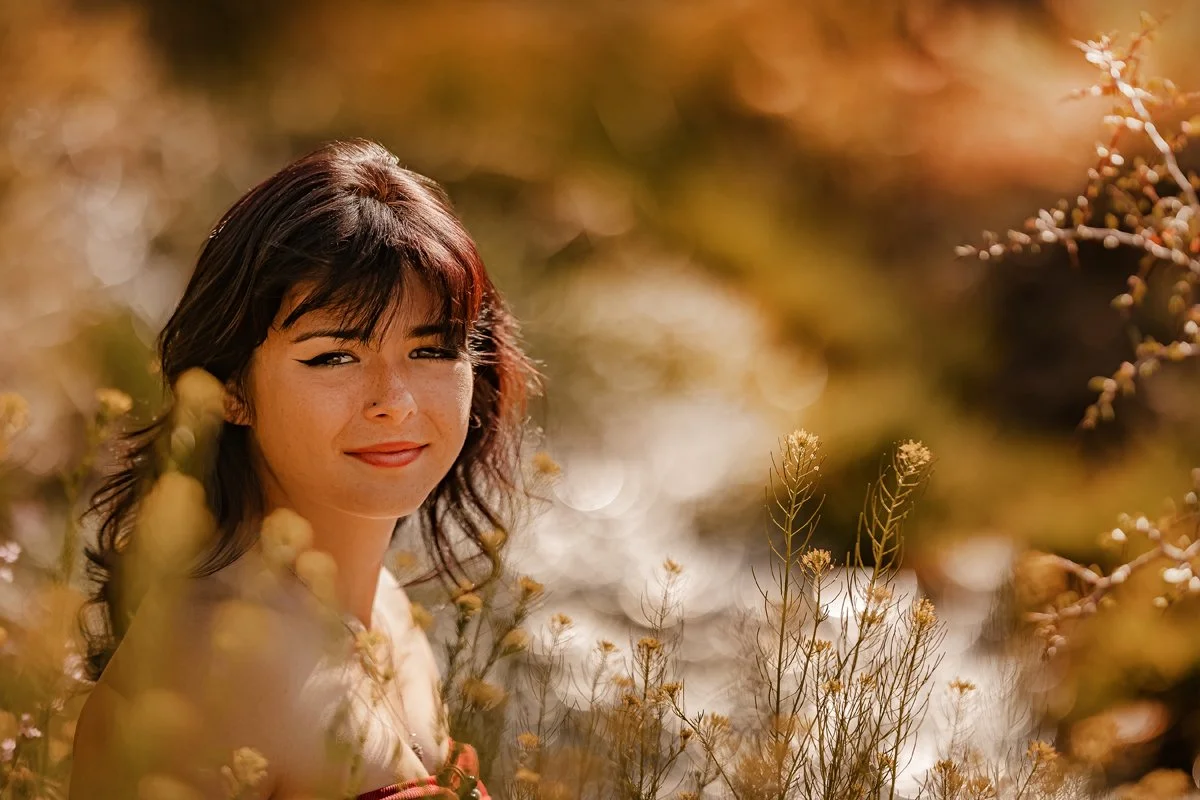 A woman with dark brown hair and freckles looking at the camera amidst golden foliage in a natural outdoor setting