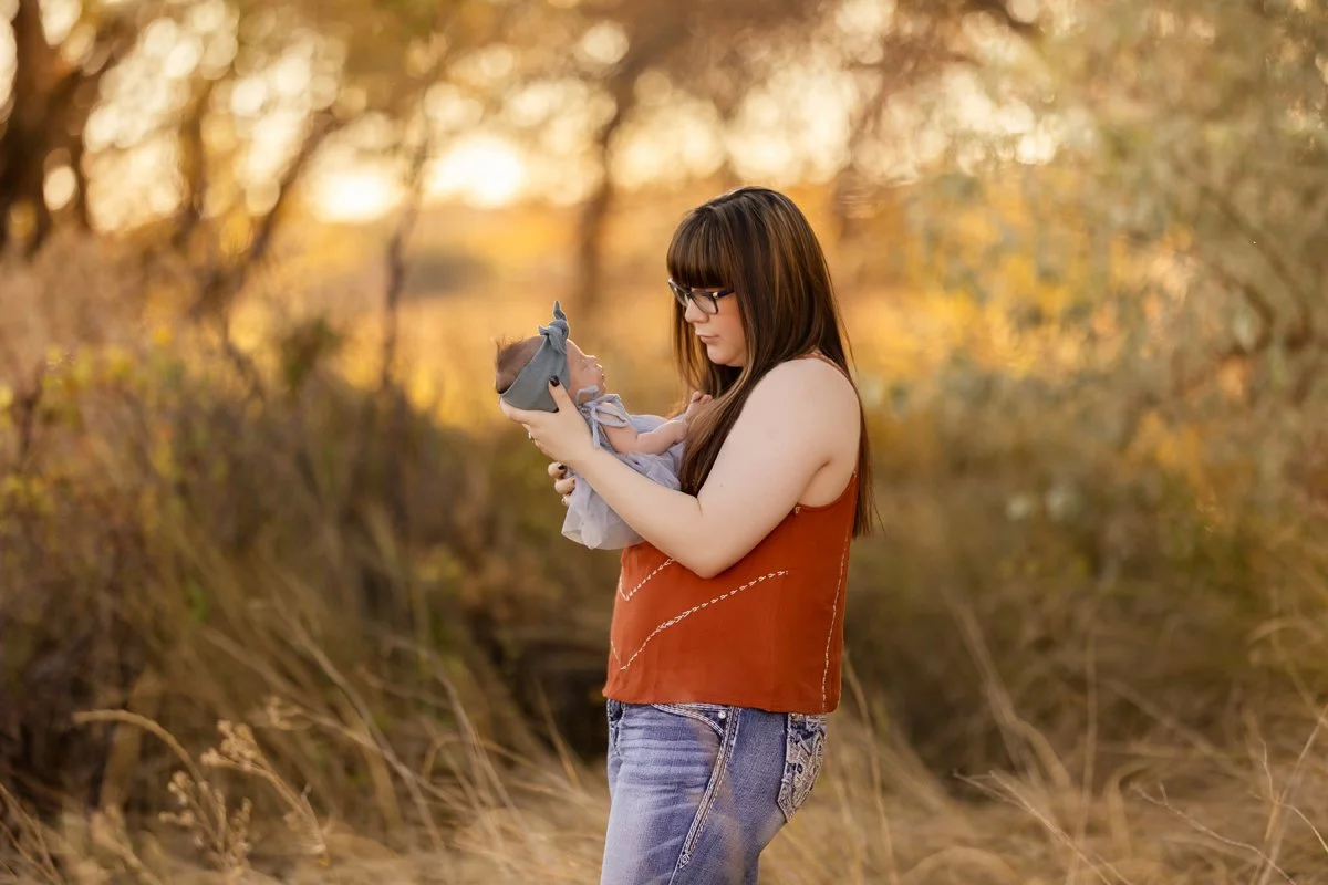 A woman with brown hair, glasses, and a sleeveless rust-colored top holding a baby wrapped in a gray blanket outdoors during autumn.