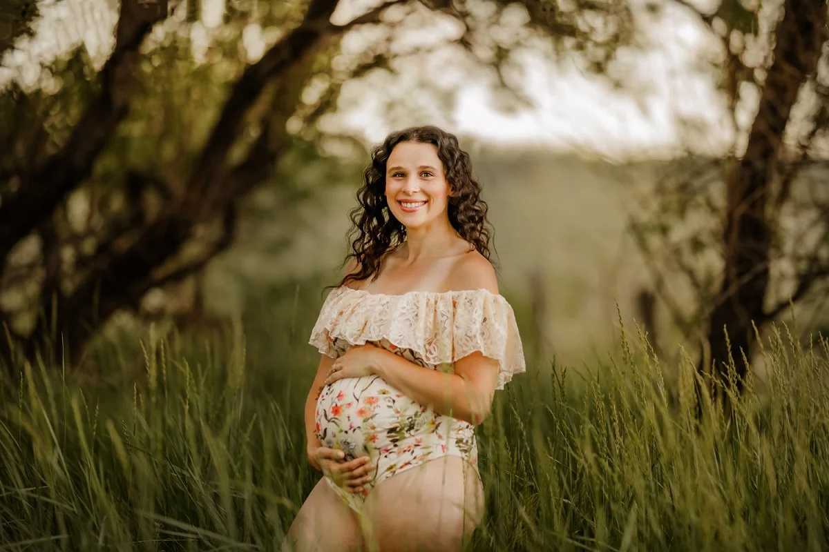 A pregnant woman smiling in a field of tall grass with trees in the background, wearing a floral off-the-shoulder dress.