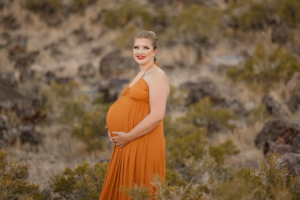 Pregnant woman standing outdoors in a desert-like area with rocks and sparse vegetation, wearing an orange dress and smiling.