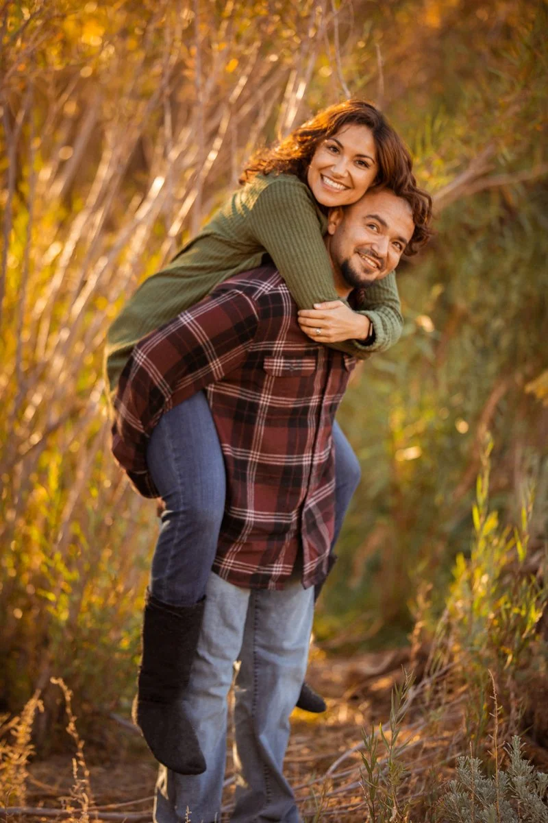 A couple enjoying a piggyback ride outdoors during fall, surrounded by golden trees and foliage.