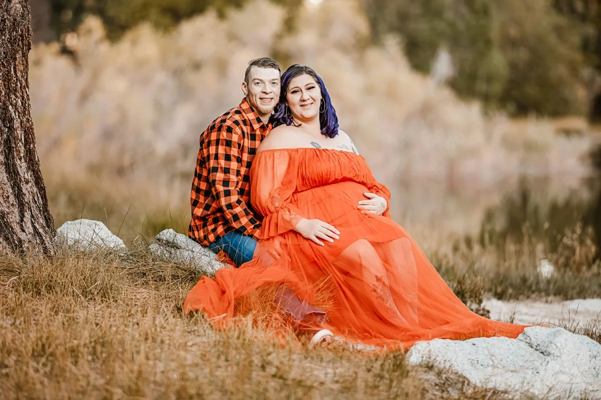 A couple sitting outdoors on rocks near a tree, with the woman pregnant, wearing a bright orange dress and the man in a red and black checkered shirt.