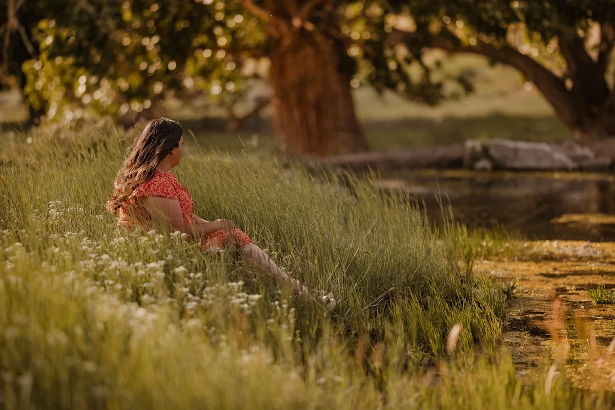 A woman with long hair sitting on a grassy riverside in a red dress, looking towards the water and trees in the background during sunset.